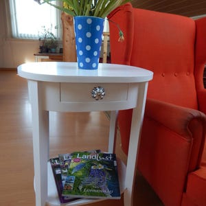 Könnte beinhalten: Ein weißer Beistelltisch mit Schublade und unterem Regal. Eine blaue Vase mit weißen Punkten enthält Blumen. Im Hintergrund ein orangefarbener Sessel. Die Schublade hat einen dekorativen Knopf.