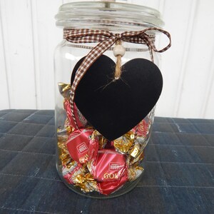 Könnte beinhalten: Ein klares Glas mit verpackten Süßigkeiten. Das Glas hat einen Deckel mit einem beigen Knopf und ist mit einem braun-weiß karierten Band und einer schwarzen herzförmigen Tafel verziert. Die Süßigkeiten sind rot und goldfarben.