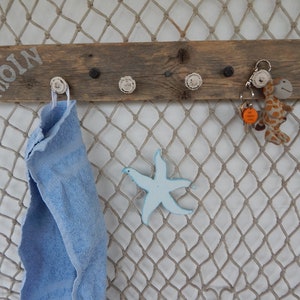 Könnte beinhalten: Ein rustikaler Wand-Kleiderhaken aus Holz mit dem Schriftzug "MOIN". Er hat mehrere Haken, ein blaues Handtuch, einen Seestern und Schlüsselanhänger mit einer Giraffe und einem orangefarbenen Anhänger. Der Hintergrund ist ein Fischernetz.