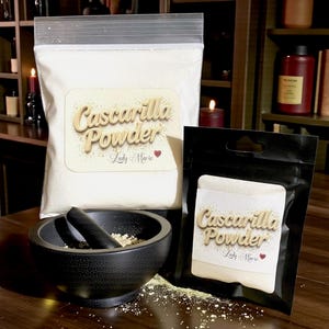 May include: A clear plastic bag and a black pouch containing white powder labeled "Cascarilla Powder" with a heart. A black mortar and pestle are in the foreground, with powder scattered around. The background includes shelves and candles.