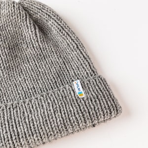 The image displays a close-up of a gray ribbed knit beanie with a folded section featuring a small, colorful logo tag. The beanie is set against a white background.