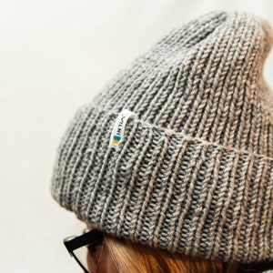The image shows a close-up of a person wearing a textured knit beanie in varying shades of gray, with a small logo on the fold.