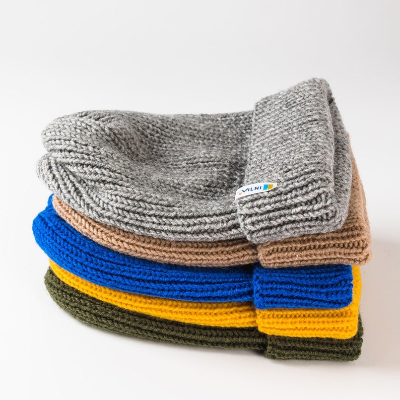 The picture showcases a stack of folded knit beanies in various colors, including gray, brown, blue, yellow, and green. The top beanie has a visible logo tag.