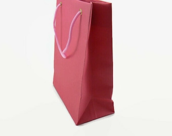 May include: A red gift bag with a pink rope handle. The bag is made of a sturdy paper material and has a rectangular shape. The handle is attached with gold-colored eyelets.