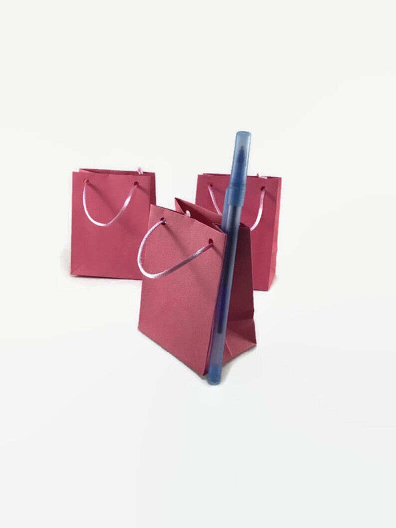 May include: Three small pink paper gift bags with white handles. A blue pen is standing upright in the center bag.