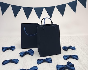 May include: Navy blue gift bags with matching bow ties and a banner. The bags have handles and are standing on a light wood surface. The bow ties are scattered around the bags. A navy blue banner is hanging in the background.