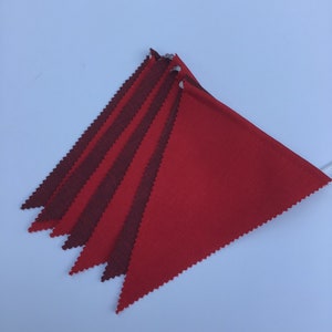 K&ouml;nnte beinhalten: Eine Girlande aus roten und kastanienbraunen Dreiecksflaggen. Die Flaggen bestehen aus einem strukturierten Stoff und sind an einer wei&szlig;en Schnur befestigt. Diagonale Anordnung. Eine festliche Dekoration.