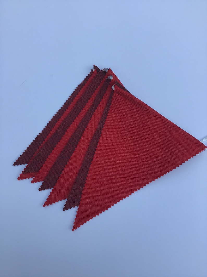 K&ouml;nnte beinhalten: Eine Girlande aus roten und kastanienbraunen Dreiecksflaggen. Die Flaggen bestehen aus einem strukturierten Stoff und sind an einer wei&szlig;en Schnur befestigt. Diagonale Anordnung. Eine festliche Dekoration.
