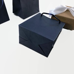 May include: Three navy blue paper gift bags with handles. Two of the bags are folded flat and one is standing upright. A small brown gift box with a white satin ribbon is in the background.