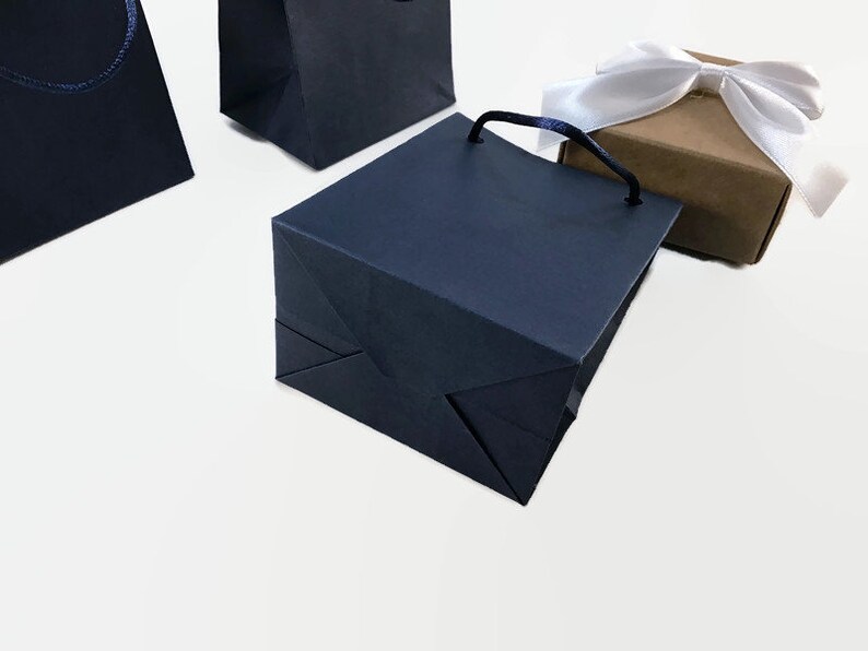 May include: Three navy blue paper gift bags with handles. Two of the bags are folded flat and one is standing upright. A small brown gift box with a white satin ribbon is in the background.