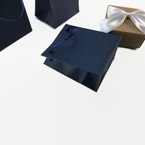May include: Three gift bags, two are navy blue with dark blue rope handles, and one is brown with a white satin bow.