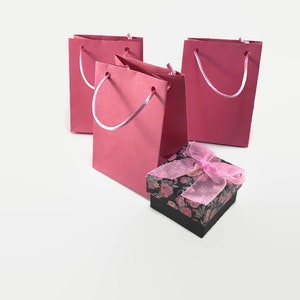 May include: Three pink paper gift bags with white handles and a small black gift box with a pink floral design and a pink ribbon.