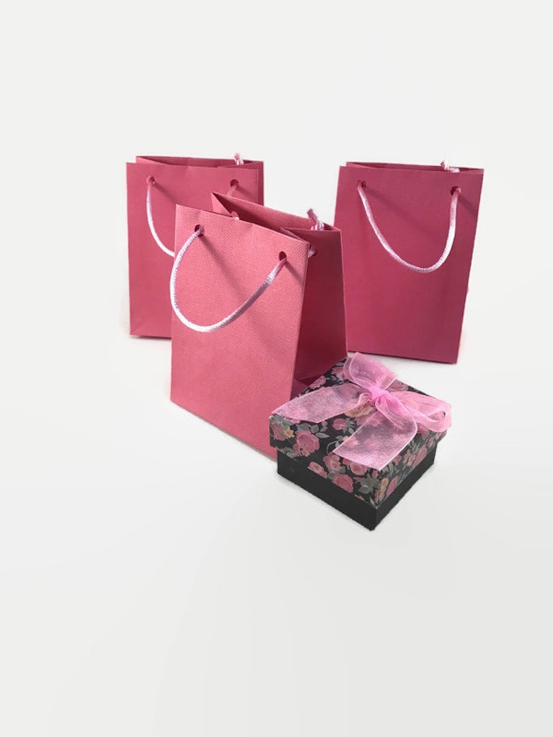 May include: Three pink paper gift bags with white handles and a small black gift box with a pink floral design and a pink ribbon.
