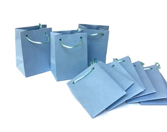 May include: A collection of small, light blue paper gift bags, each featuring thin, mint green ribbon handles threaded through punched holes. Three bags stand upright, while several others are folded flat and stacked together.