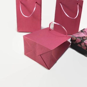May include: Three pink paper gift bags with white ribbon handles. The bags are on a white surface.