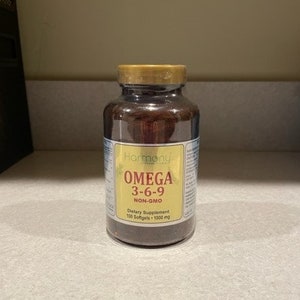 May include: A brown glass bottle with a gold lid and label. The label reads "Harmony" and "Omega 3-6-9 Non-GMO Dietary Supplement 100 Softgels - 1000 mg".
