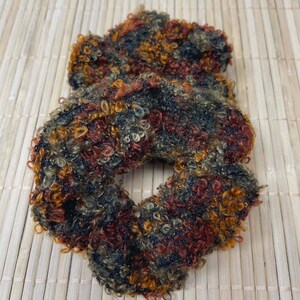 Könnte beinhalten: Ein mehrfarbiges Scrunchie mit einem strukturierten, geschlungenen Design. Das Scrunchie hat eine Mischung aus dunkelroten, orangefarbenen, schwarzen und olivgrünen Garnen. Das Haaraccessoire ist aus einem weichen Material, möglicherweise Wolle.