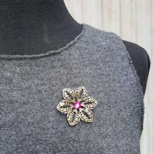 Könnte beinhalten: Eine goldfarbene Blumenbrosche mit einem rosafarbenen Strassstein in der Mitte. Die Brosche befindet sich auf einem grauen Pullover.