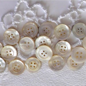 Peut inclure: Un ensemble de 15 boutons en nacre blanche à quatre trous chacun. Les boutons sont disposés sur une dentelle blanche au crochet.