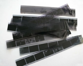 May include: A collection of black and gray 35mm film strips, each with multiple frames. The film strips are scattered across a white surface, showing various images. The film has sprocket holes along the edges.