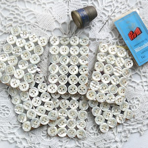 Puede incluir: Un surtido de suministros de costura vintage, que incluye un dedal, agujas y un paquete de agujas etiquetado como "Prym". Varias tarjetas de botones blancos de cuatro agujeros están dispuestas sobre una superficie de encaje blanco, mostrando un tema artesanal clásico.