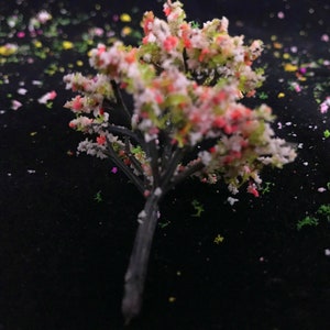 May include: A small, artificial tree with pink and white flowers and a black trunk. The tree is on a black background with small, colorful confetti.