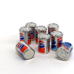May include: Eight miniature silver cans of soda pop with red, white, and blue labels. The cans are arranged in a circle on a white surface.