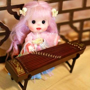 Könnte beinhalten: Eine Puppe mit pinkfarbenem Haar spielt eine braune, hölzerne Guzheng, ein traditionelles chinesisches Musikinstrument mit Saiten.
