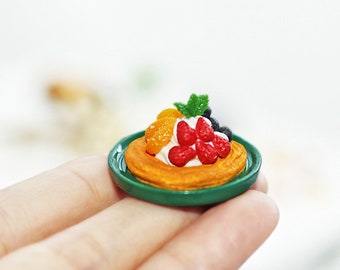 Könnte beinhalten: Ein Miniaturgebäck mit einer Cremefüllung, belegt mit Erdbeeren, Orangenscheiben und einem grünen Blatt auf einem kleinen grünen Teller.