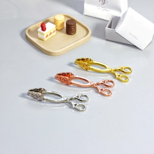 May include: Three pairs of decorative tongs in gold, silver, and copper finishes. The tongs have a delicate, ornate design and are perfect for serving small items.