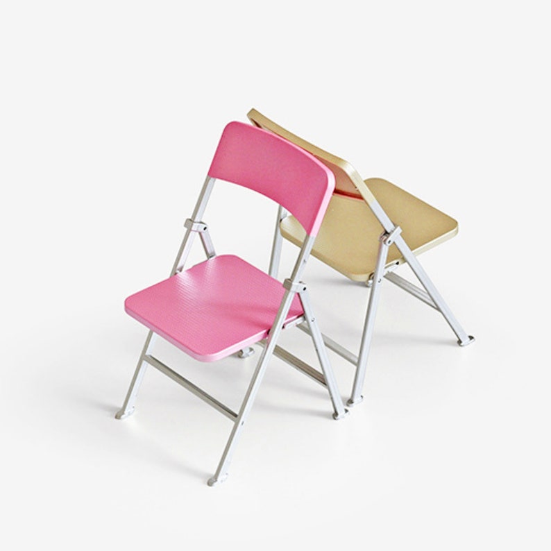 May include: Two folding chairs, one pink and one beige, are stacked on top of each other. The chairs have a metal frame and a wooden seat.