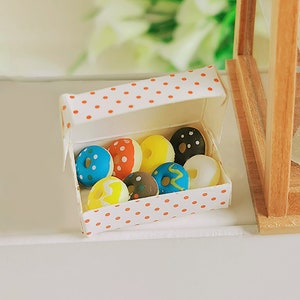 May include: A white box with red polka dots, open to reveal a variety of colorful donuts with different designs. The donuts are arranged in a single layer inside the box.