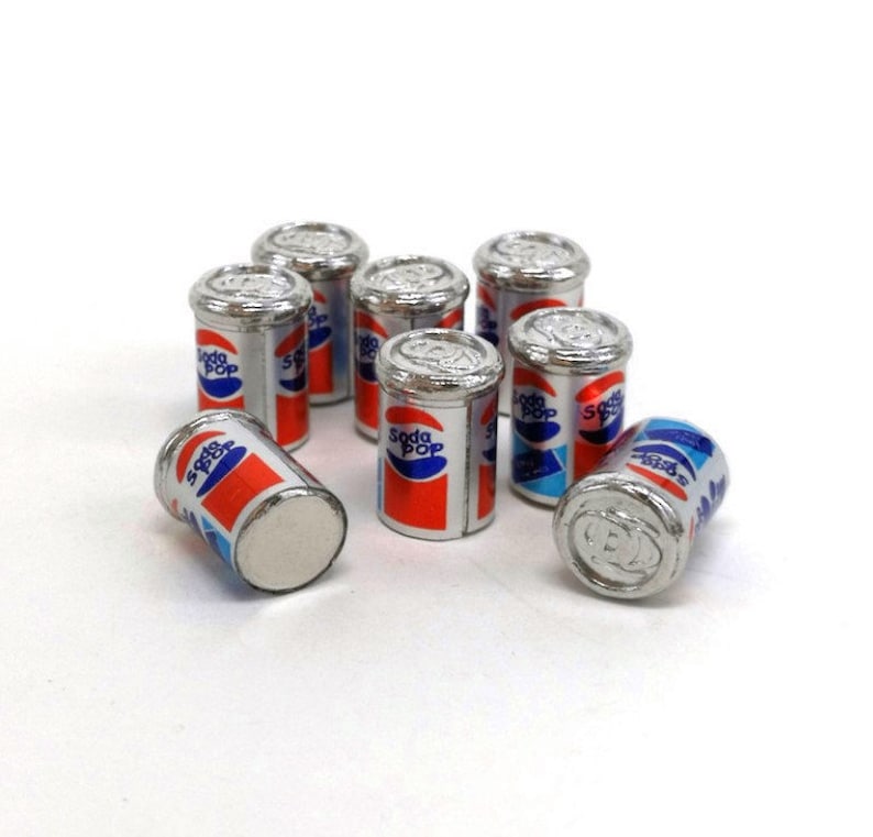 May include: Eight miniature silver cans of soda pop with red, white, and blue labels. The cans are arranged in a circle on a white surface.