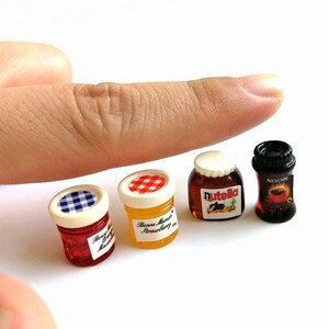 May include: Four miniature glass jars with lids. The jars contain red, yellow, brown, and black substances. The jars are labeled "Bonne Maman Strawberry", "Nutella", and "Nescafe".