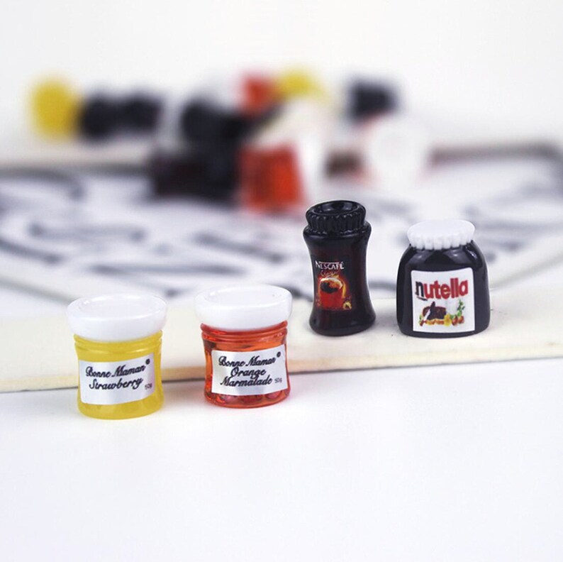 May include: Four miniature glass jars with white lids. The jars contain orange marmalade, strawberry jam, Nutella, and Nescafe coffee. The jars are on a white surface.
