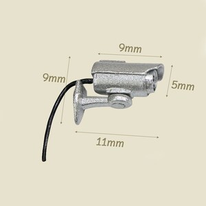 K&ouml;nnte beinhalten: Eine silberne Miniatur-&Uuml;berwachungskamera mit einem schwarzen Kabel, die 9 mm hoch, 11 mm breit und 5 mm tief ist.