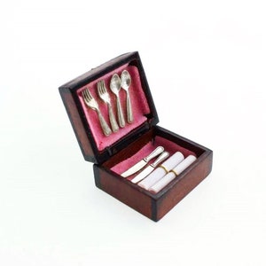 Puede incluir: Una caja de madera con un forro de terciopelo rosa, que contiene un juego de cubiertos de plata en miniatura y servilletas enrolladas. La caja está abierta y los cubiertos están ordenados cuidadosamente en su interior.