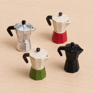 Puede incluir: Cuatro cafeteras moka en miniatura de diferentes colores. Una es de color plata metálico, una tiene la parte superior crema con base roja, una tiene la parte superior crema con base verde y una es negra. Cada una tiene un asa negra y un pomo.