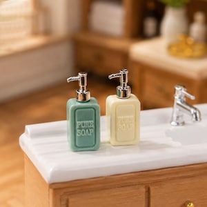 May include: Two miniature soap dispensers, one pale green and one cream-colored, sit side-by-side on a white countertop. Each dispenser has a silver pump and the words "PURE SOAP" are printed on the front. A miniature faucet is visible in the background.