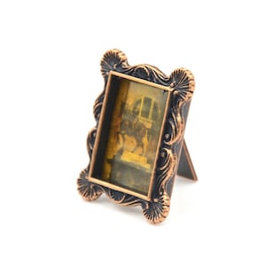 May include: Small, bronze-toned picture frame with an ornate, scalloped design. The frame holds a vintage-style image, likely a photograph, with sepia tones. It features a built-in easel back for standing display. The frame is made of metal.