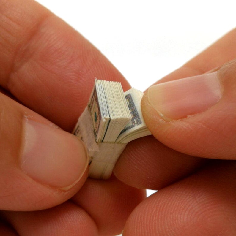 May include: A stack of US dollar bills folded and held in a hand. The bills are folded in half and then rolled up tightly. The person is holding the rolled up bills in their hand, with their fingers wrapped around the bills.