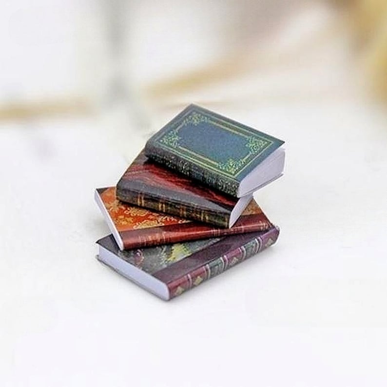 May include: A stack of four miniature books with different colored covers. The top book has a green cover with gold accents, the second book has a red cover with gold accents, the third book has a brown cover with gold accents, and the bottom book has a dark blue cover with gold accents.
