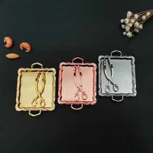 Puede incluir: Tres bandejas de servir decorativas con pinzas a juego. Las bandejas son cuadradas y tienen un borde elevado. Las bandejas son doradas, doradas rosadas y plateadas.