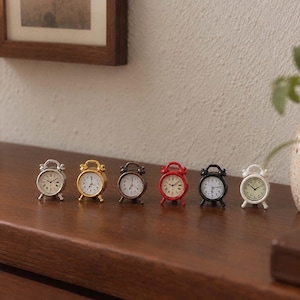 May include: A row of six miniature alarm clocks in silver, gold, bronze, red, black, and white finishes. Each clock features a round face with black numerals, two small bells on top, and two legs, standing in a line on a wooden surface.