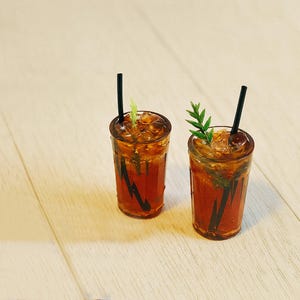 Könnte beinhalten: Zwei Miniatur-Eistees in klaren Gläsern mit schwarzen Strohhalmen. Jedes Getränk ist mit Eiswürfeln gefüllt und mit einem Zweig Grünzeug garniert. Die Gläser haben eine dunkelbraune Flüssigkeit und schwarze dekorative Akzente.