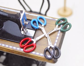 May include: Four miniature scissors with different colored handles: red, blue, green, and black. The scissors are arranged in a cross pattern on a wooden surface.