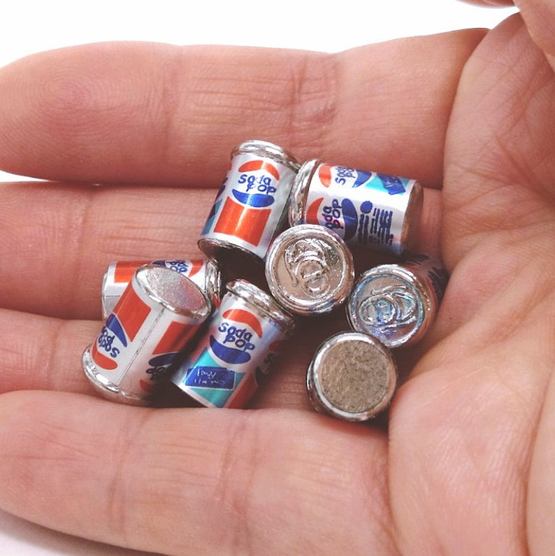 May include: A collection of miniature silver soda pop cans with blue and red labels. The cans have the words "Soda Pop" printed on them.
