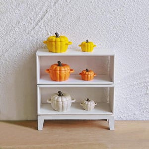 Può includere: Una mensola bianca a tre livelli espone sei contenitori in miniatura a forma di zucca. Le zucche sono gialle, arancioni e bianche, ognuna con un gambo marrone scuro. La mensola è contro un muro bianco strutturato, creando un'atmosfera autunnale.
