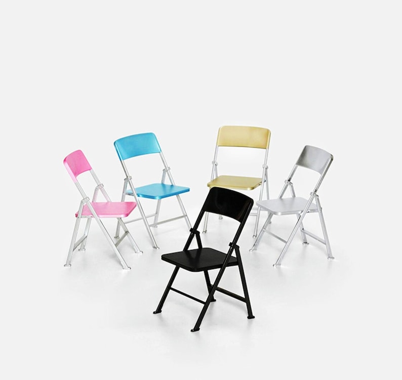 May include: Five folding chairs in different colors: pink, blue, gold, silver, and black. The chairs are arranged in a circle on a white background.