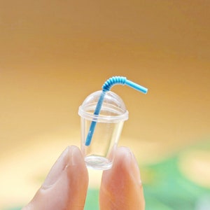 Puede incluir: Un vaso de pl&aacute;stico transparente en miniatura con una pajita azul. El vaso tiene una tapa y se sostiene entre dos dedos.